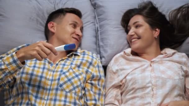 мужчина и женщина целуются, лежа в постели и показывая положительный тест на беременность, размытый, вид сверху - Кадры, видео