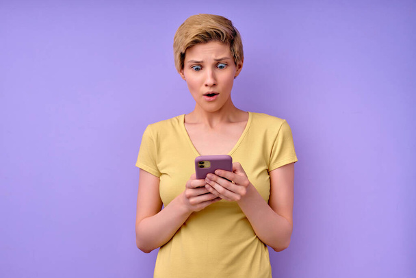 Shocked young woman in casual t-shirt, keeping mouth widely opened,looking at smartphone - Photo, Image