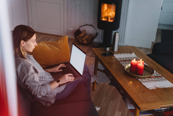 Side view of chilling woman on couch near warm fireplace typing on laptop with blank screen spending time at home - Valokuva, kuva