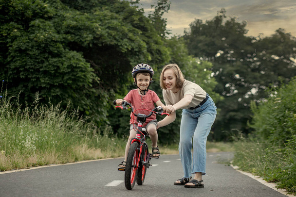 Mother teaching son to ride bicycle. Happy cute boy in helmet learn to riding a bike on the bike path in summer raining day at sunset time. Family weekend. - Foto, immagini