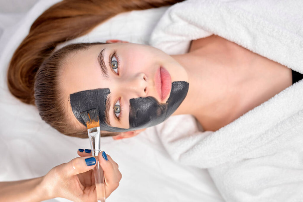 Woman relaxing with charcoal facial mask, cropped professional beautician applying on face - Zdjęcie, obraz