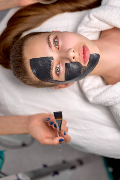 Top view on woman with black cosmetic facial mask lying on bed at spa. Face peeling - Zdjęcie, obraz