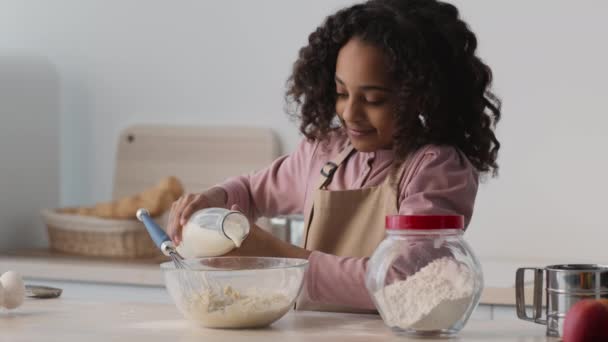 Pequeña chica afroamericana rizada agregando leche a otros ingredientes, preparando masa para pastel dulce en la cocina - Imágenes, Vídeo