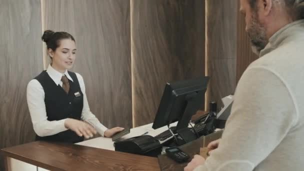 Medium shot of mature businessman paying for accommodation in modern hotel by credit card check-in at reception desk. Женщина-секретарша, стоящая у стойки - Кадры, видео