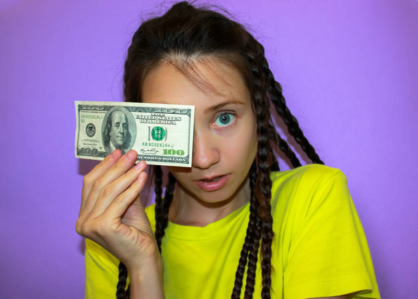 Surprised brunette blue-eyed girl in a yellow T-shirt on a purple background holding a hundred dollar note in hand, covering face. Success, business investment, finance, earning money, cash concept. - Foto, immagini