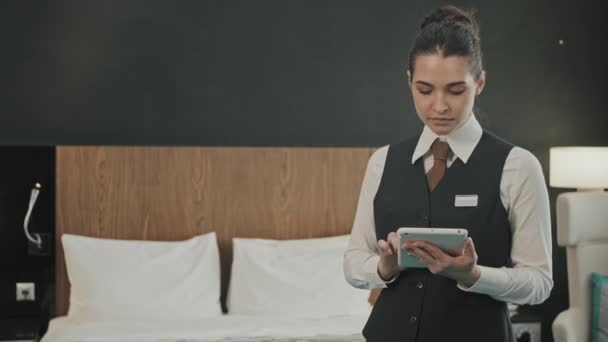 Retrato panorámico medio con lentitud de la gerente de hotel joven en uniforme usando tableta digital de pie en la habitación de hotel cómoda moderna y luego mirando a la cámara - Metraje, vídeo