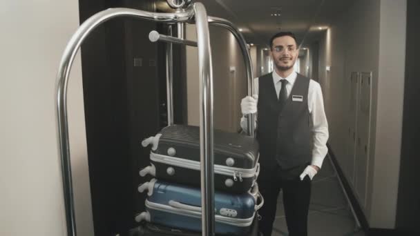 Retrato de tiro de arco médio com lentidão de porteiro masculino profissional em luvas e uniforme posando para câmera com carrinho de bagagem no corredor do hotel - Filmagem, Vídeo