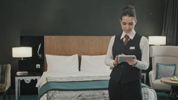 Portrait slow moyen de la jeune hôtelière en uniforme souriant à l'appareil photo avec tablette numérique dans les mains debout dans une chambre d'hôtel moderne et confortable - Séquence, vidéo
