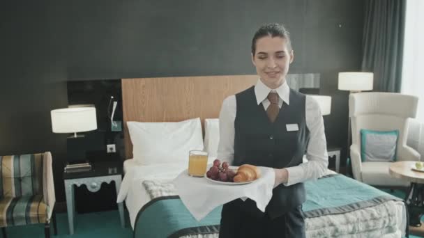 Mittelschweres Porträt einer jungen Kellnerin, die mit frischem Frühstück auf Tablett neben einem King-Size-Bett im modernen Hotelzimmer für die Kamera posiert - Filmmaterial, Video