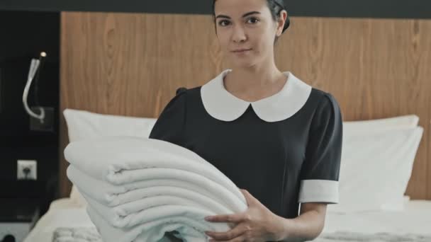 Tilting-up portrait with slowmo of young smiling woman working as housekeeper in hotel holding white towels standing next to king-size bed in hotel room and posing for camera - Footage, Video
