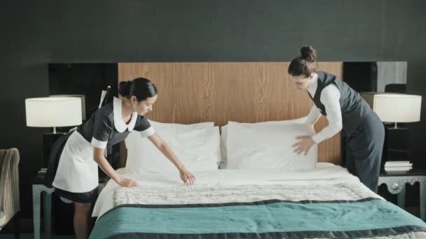 Mediana toma de gerente de hotel femenino y ama de llaves en uniforme haciendo la cama juntos en la habitación de los huéspedes, preparándose para su llegada - Imágenes, Vídeo