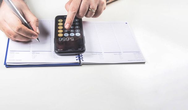 Hombre haciendo cálculos con su calculadora de teléfono móvil y escribiendo en un cuaderno de encuadernación con un bolígrafo. Presupuesto y economía. - Foto, Imagen