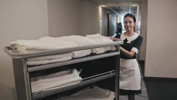 Retrato de cámara lenta zoom de una joven ama de llaves en uniforme blanco y negro sonriendo a la cámara de pie con toallas blancas y ropa de cama en el carrito de la mano en el pasillo del hotel - Imágenes, Vídeo