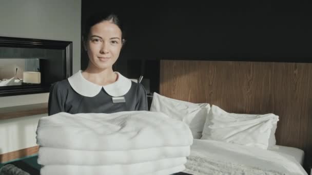 Retrato medio de la joven ama de llaves sonriendo a la cámara mientras sostiene toallas blancas frescas de pie en la habitación de hotel de lujo - Imágenes, Vídeo