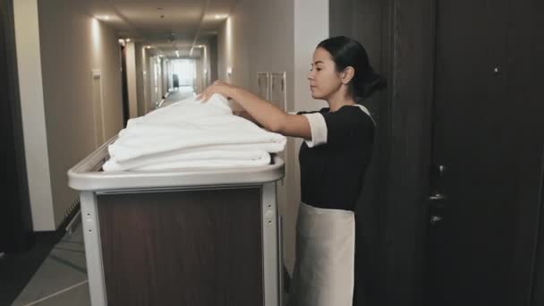 Μεσαία βολή της γυναίκας οικονόμου με πετσέτες, λινά και προμήθειες καθαρισμού στο καλάθι χέρι με τα πόδια κατά μήκος μακρύ διάδρομο, την είσοδο δωμάτιο του ξενοδοχείου για τον καθαρισμό πόρτα ανοίγματος με κλειδί κάρτας - Πλάνα, βίντεο