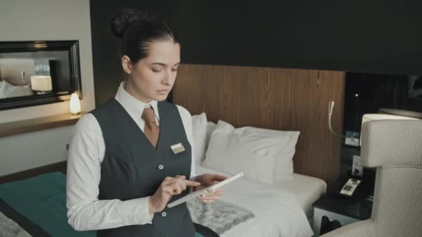 Střední pomalý portrét usměvavé ženy hotelového manažera pomocí digitálního tabletu, pak pohled na kameru stojící v interiéru moderní high-class hotelový pokoj - Záběry, video