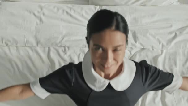 Slowmo shot of relaxed female housekeeper lying down on her back on luxury hotel bed with white linens with arms spread resting after work - Footage, Video
