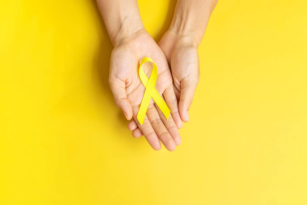 hand holding Yellow Ribbon on yellow background for supporting people living and illness. September Suicide prevention day, Childhood Cancer Awareness month and World cancer day concept - Zdjęcie, obraz