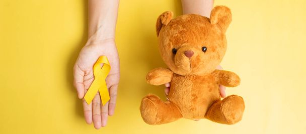 mano que sostiene la cinta amarilla y la muñeca del oso en el fondo amarillo para apoyar a niños que viven y se enferman. Septiembre Concepto de mes de concienciación sobre el cáncer infantil y día mundial del cáncer - Foto, Imagen