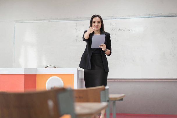 Foto d'archivio ritratto di una donna asiatica allegra e sicura di sé insegnante in uniforme tuta nera con tablet digitale e laptop per insegnare la lingua moderna in classe - Foto, immagini