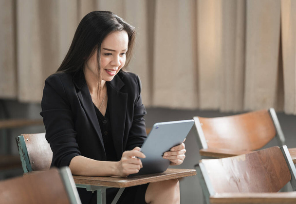 Foto retrato de stock de una mujer asiática alegre y confiada maestra en un traje de negocios negro uniforme con una tableta digital y un ordenador portátil para enseñar el idioma moderno en el aula - Foto, imagen