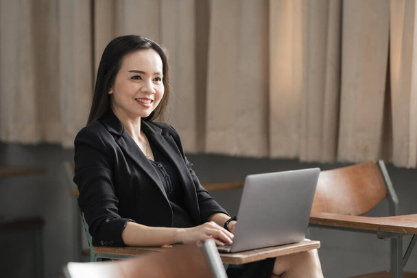 Stock photo portrait of a confident cheerful Asian woman teacher in a black business suit uniform with a digital tablet and laptop to teach modern language in the classroom - Zdjęcie, obraz