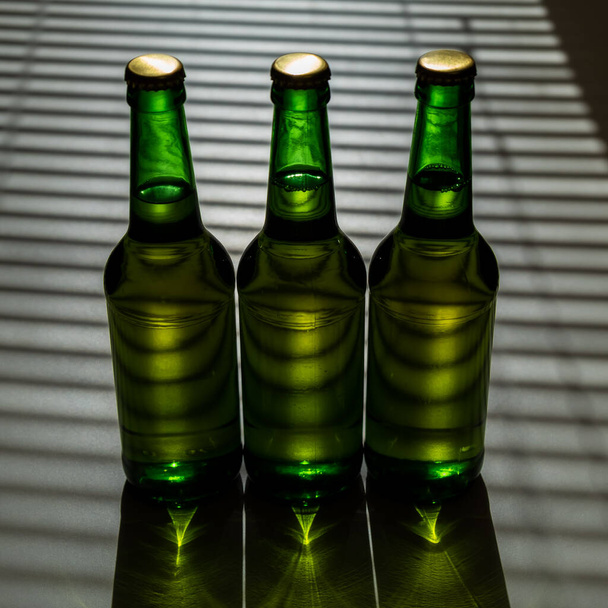 Three green glass beer bottles in the shade of the blinds. - Photo, Image