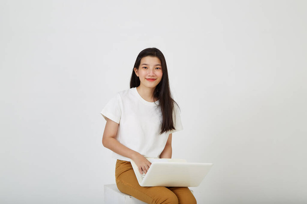 Счастливая привлекательная молодая женщина с ноутбуком сидя скрестив ноги и глядя на пустое пространство на белом фоне студии - Фото, изображение