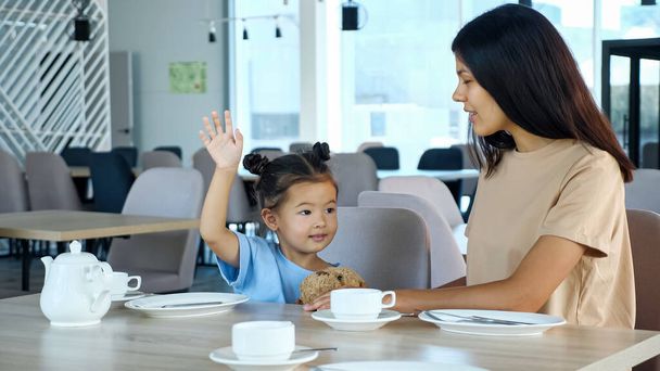 Maluch dziewczyna podnosi rękę z uśmiechem siedzi przez mamę w kawiarni - Zdjęcie, obraz