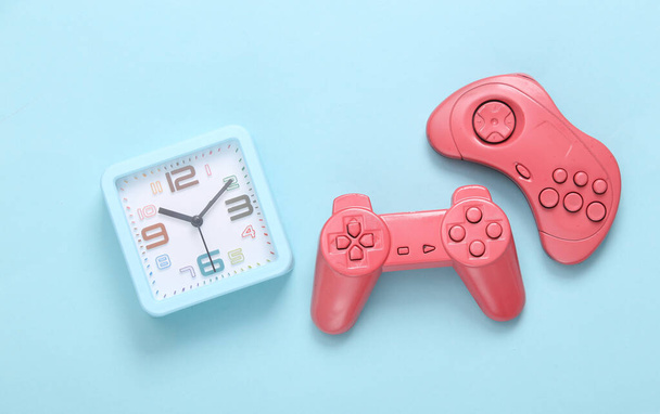 Два рожевих геймпади з будильником на синьому фоні. Час грати у відеоігри
 - Фото, зображення