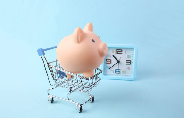 Piggy bank in shopping trolley and alarm clock on blue background - Photo, Image