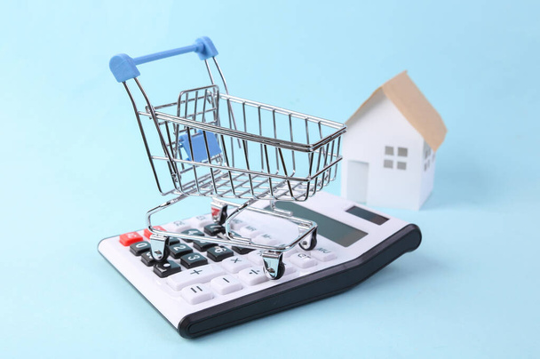 Carrito de compras con calculadora y casa modelo sobre fondo azul. Concepto de compra - Foto, imagen