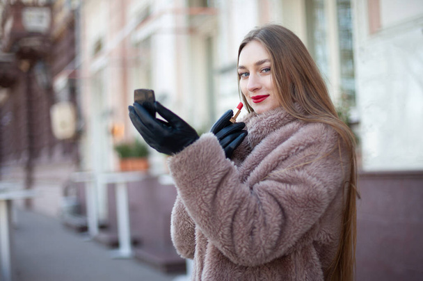 Luxury attractive woman wearing a fur coat paints her lips with red lipstick in city. Makeup. - Foto, Imagem