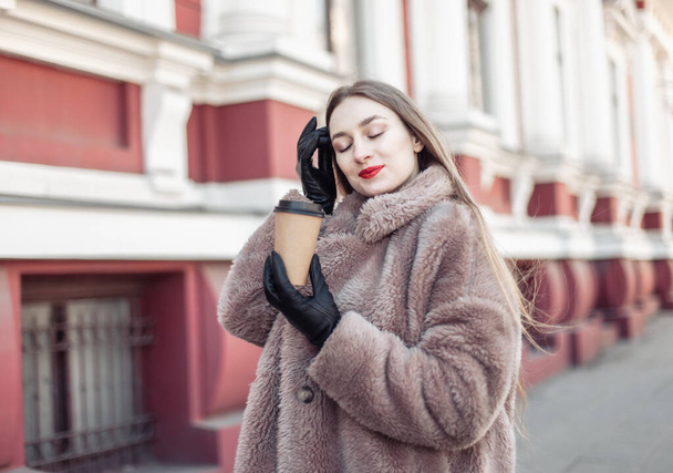Jeune femme de mode en manteau de fourrure luxueux boit du café sur la route dans la ville - Photo, image