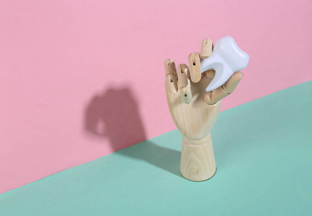 Wooden hand holding tooth on blue pink pastel background. Trendy shadow. Concept art. Minimalism. Creative layout - Foto, imagen