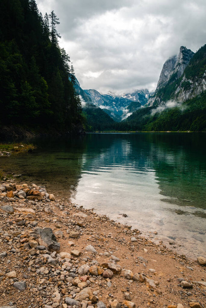 A vertical shot of Lake Gosausee in Austria with hazy reflections of green hills under a gray cloudy sky - Foto, Imagem