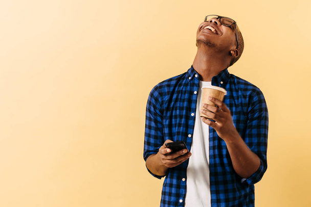 Ein junger afroamerikanischer Mann hält eine Tasse Kaffee und ein Telefon und lacht. - Foto, Bild