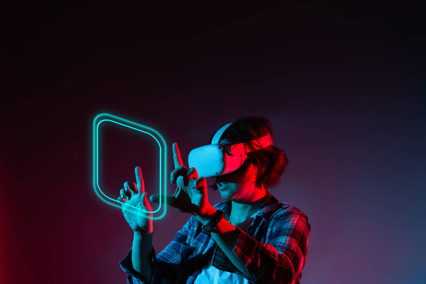 Caucasian woman 35-40 years in vr glasses, using virtual reality headset, trying to touch something with hand in neon light. - Φωτογραφία, εικόνα