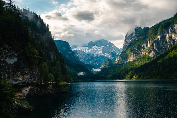 A gorgeous day at Lake Gosausee in Dachstein, Austria with tree-covered cliffs around under a bright cloudy sky - 写真・画像