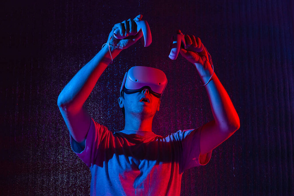 Caucasian man 25-30 years in vr glasses, playing video games with virtual reality headset, trying to touch something with hand in neon light. - Zdjęcie, obraz