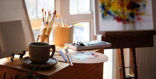 Vista trasera de manos de artista femenina sosteniendo pinceles y paleta mientras se sienta frente a la pintura de lienzo en un estudio de color blanco brillante. - Foto, imagen