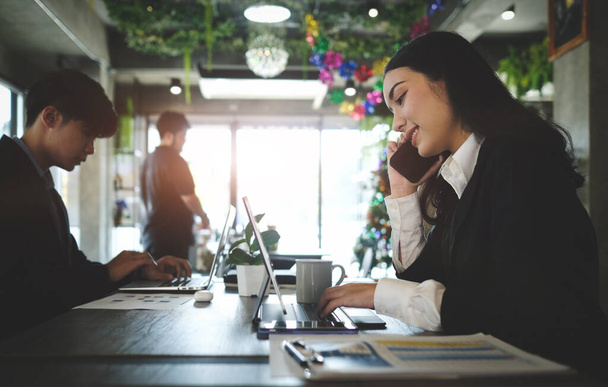 Młoda bizneswoman w czarnym garniturze rozmawiająca przez telefon komórkowy i pracująca z tabletem komputerowym siedząc z kolegą w nowoczesnym miejscu pracy. - Zdjęcie, obraz