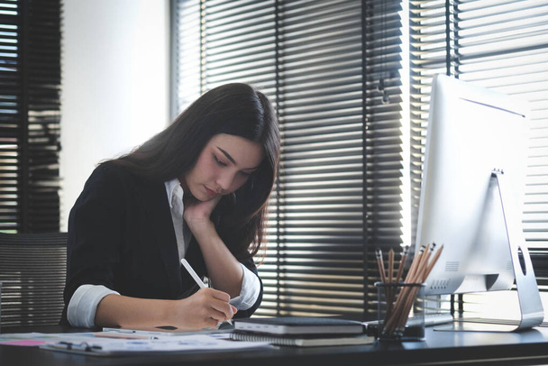 現代的なオフィスに座っている間、コンピュータと手でノートブックにメモを取りながら作業黒のスーツの幸せなビジネス女性. - 写真・画像