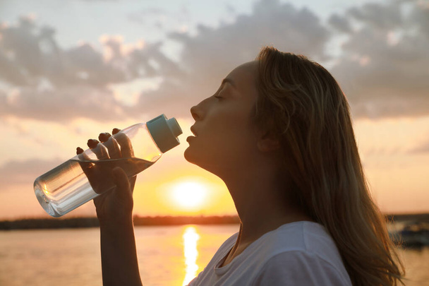 Mladá žena pitná voda, aby se zabránilo úder tepla v blízkosti řeky při západu slunce - Fotografie, Obrázek