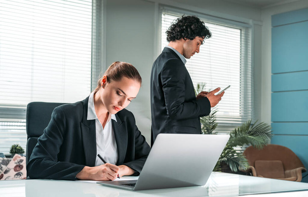 Two working concentrated business people, one is sitting at the desk in front of laptop, second is using smartphone at office background. Concept of teamwork - 写真・画像
