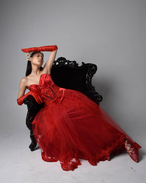 Portret pięknej młodej Azjatki w czerwonym gorsecie, długich rękawiczkach operowych i ozdobnej gotyckiej koronie królowej. Graceful pozowanie podczas siedzenia na krześle, izolowane na tle studio. - Zdjęcie, obraz