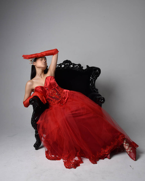 Full length  portrait of beautiful young asian woman wearing red corset, long opera gloves and ornate gothic queen crown. Graceful posing while seated on chair, isolated on studio background. - Φωτογραφία, εικόνα