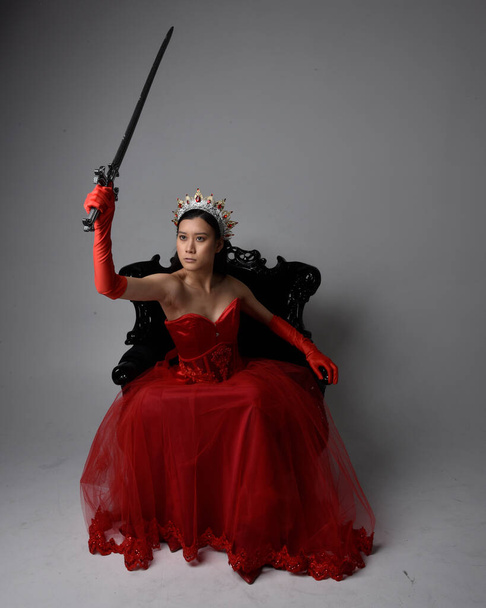 Πλήρες πορτραίτο μιας όμορφης νεαρής Ασιάτισσας με κόκκινο κορσέ, μακριά γάντια όπερας και περίτεχνο γοτθικό στέμμα βασίλισσας. Μακάριοι θέτουν, ενώ κάθεται στην καρέκλα, απομονώνονται σε στούντιο φόντο. - Φωτογραφία, εικόνα