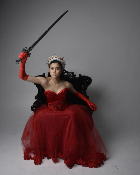 Täyspitkä muotokuva kaunis nuori aasialainen nainen yllään punainen korsetti, pitkä ooppera käsineet ja koristeellinen goottilainen kuningatar kruunu. Graceful poseeraa istuessaan tuolilla, eristetty studion taustalla. - Valokuva, kuva