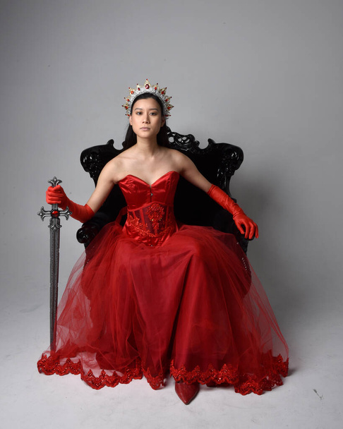 Retrato de larga duración de una hermosa joven asiática con corsé rojo, guantes largos de ópera y corona de reina gótica adornada. Posando elegante mientras está sentado en la silla, aislado en el fondo del estudio. - Foto, imagen